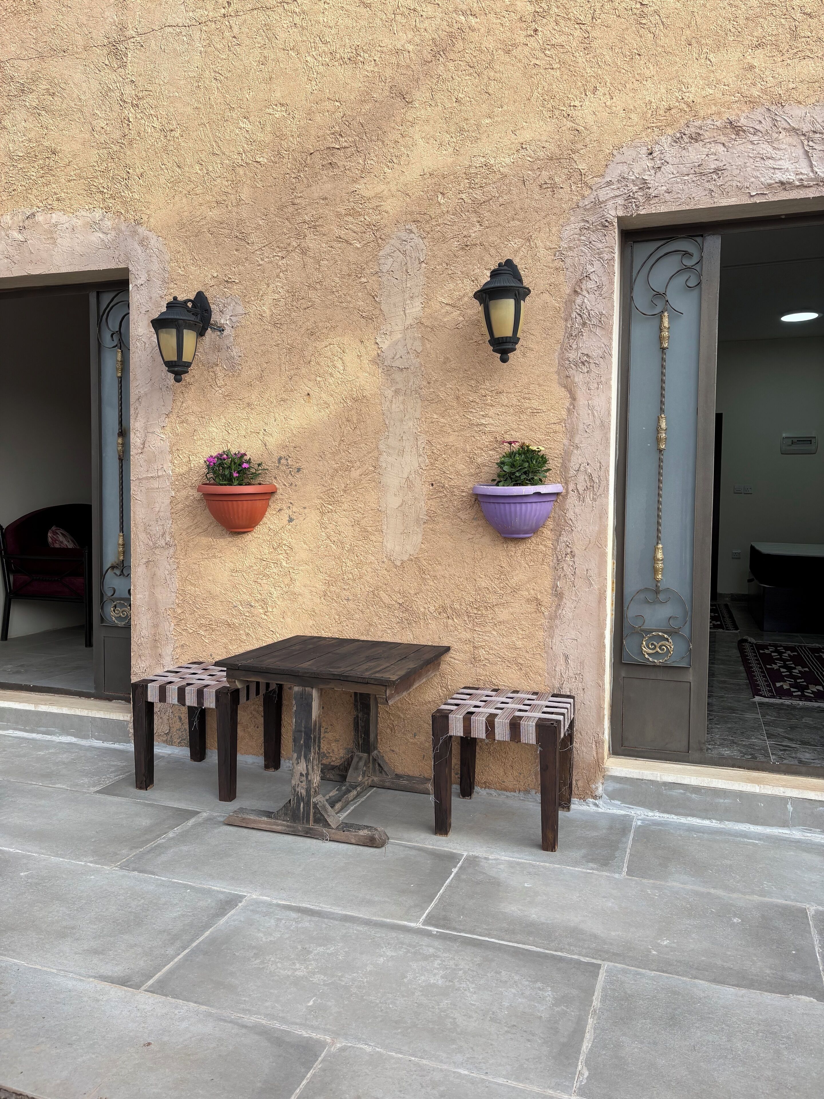 An outdoor seating area with a table and two chairs, flanked by two potted plants and two wall-mounted lamps.