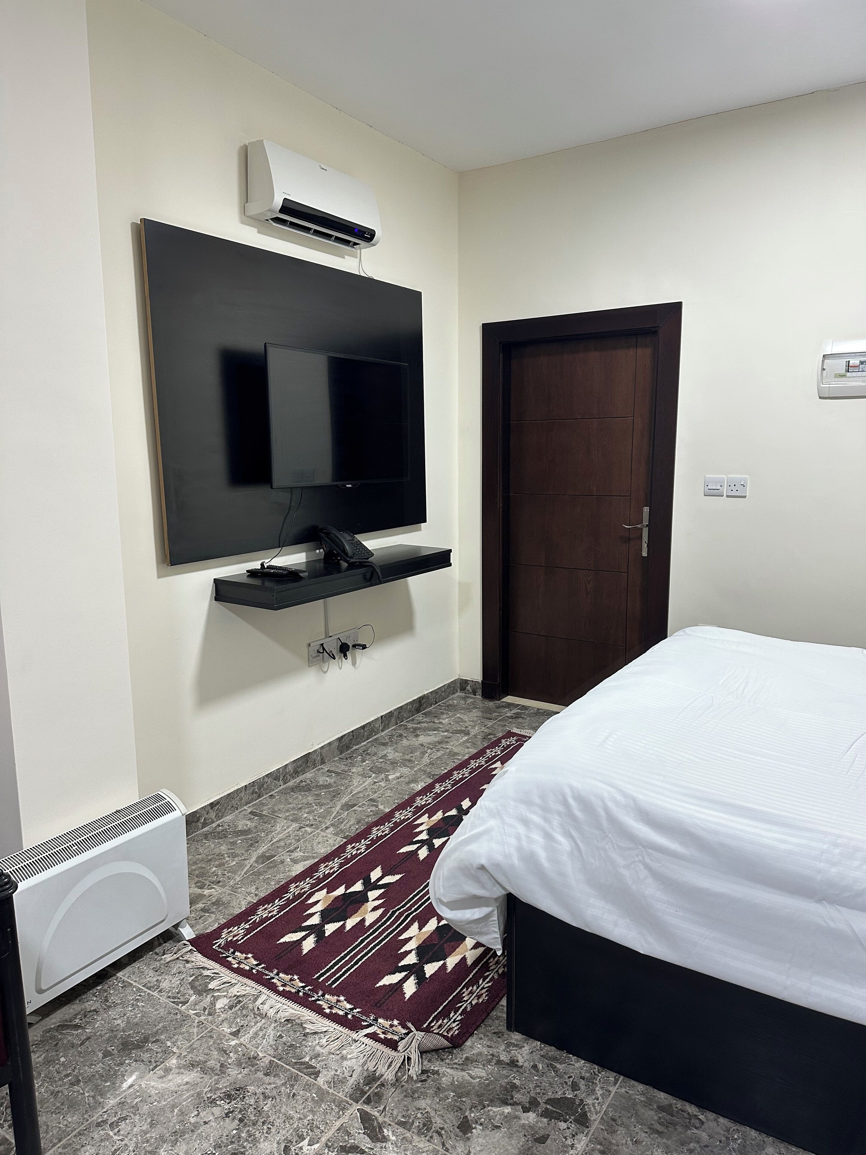 Une chambre à coucher avec un lit, un téléviseur fixé au mur, un radiateur, une porte et un tapis à motifs.