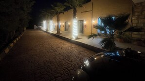A nighttime street view with a paved road, palm trees, and a building with illuminated entryways. - Qastal Zaman (Amman)