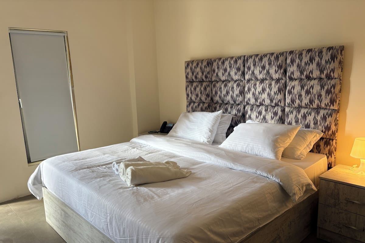 A neatly made bed with white linens and pillows, a patterned headboard, a bedside phone, and a nightstand with a lamp.