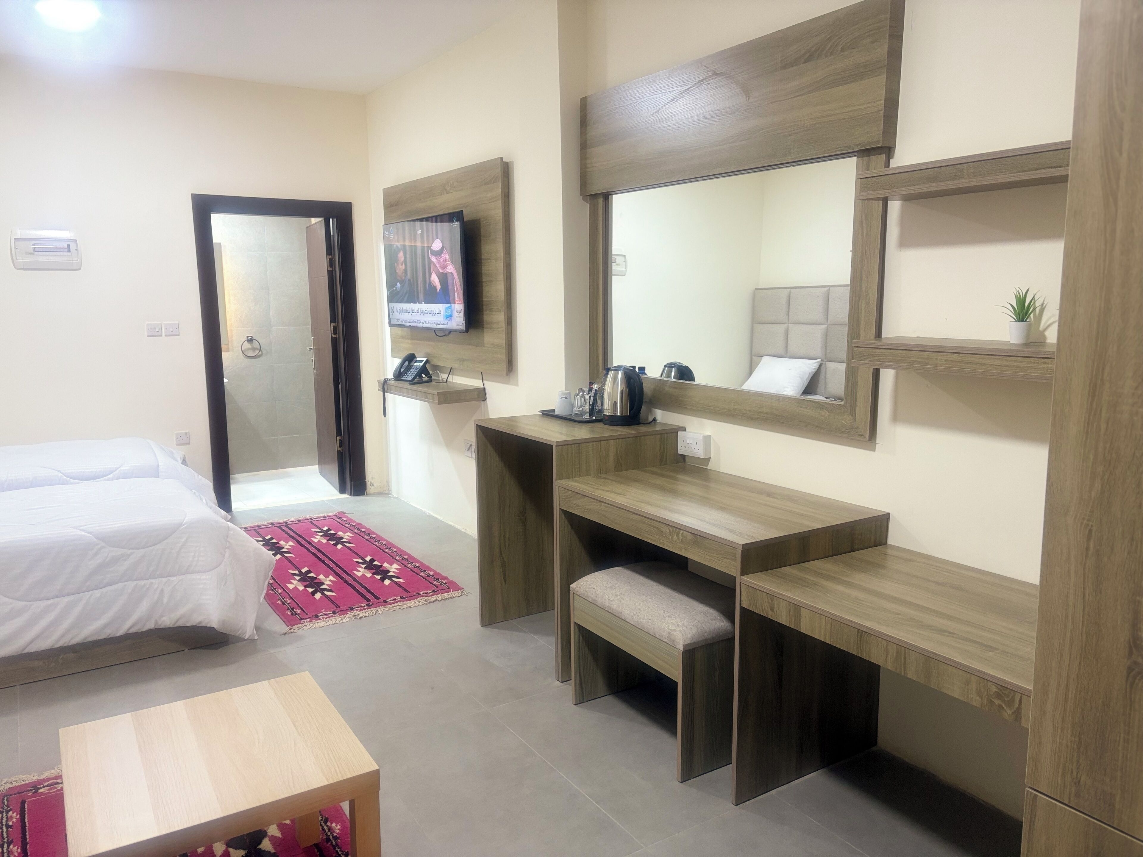 A hotel room with a bed, a desk, a chair, a small table, a mirror, a TV, and a shelf with a plant.