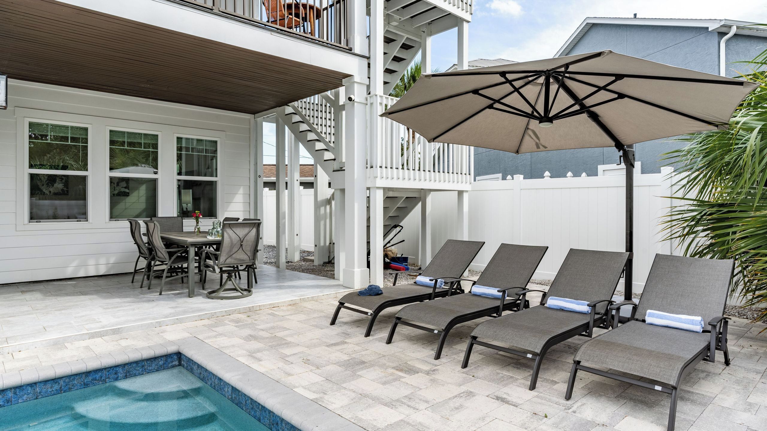 Un área junto a la piscina con una mesa y sillas, una zona de estar cubierta con sillones de descanso y una escalera que conduce a una terraza.