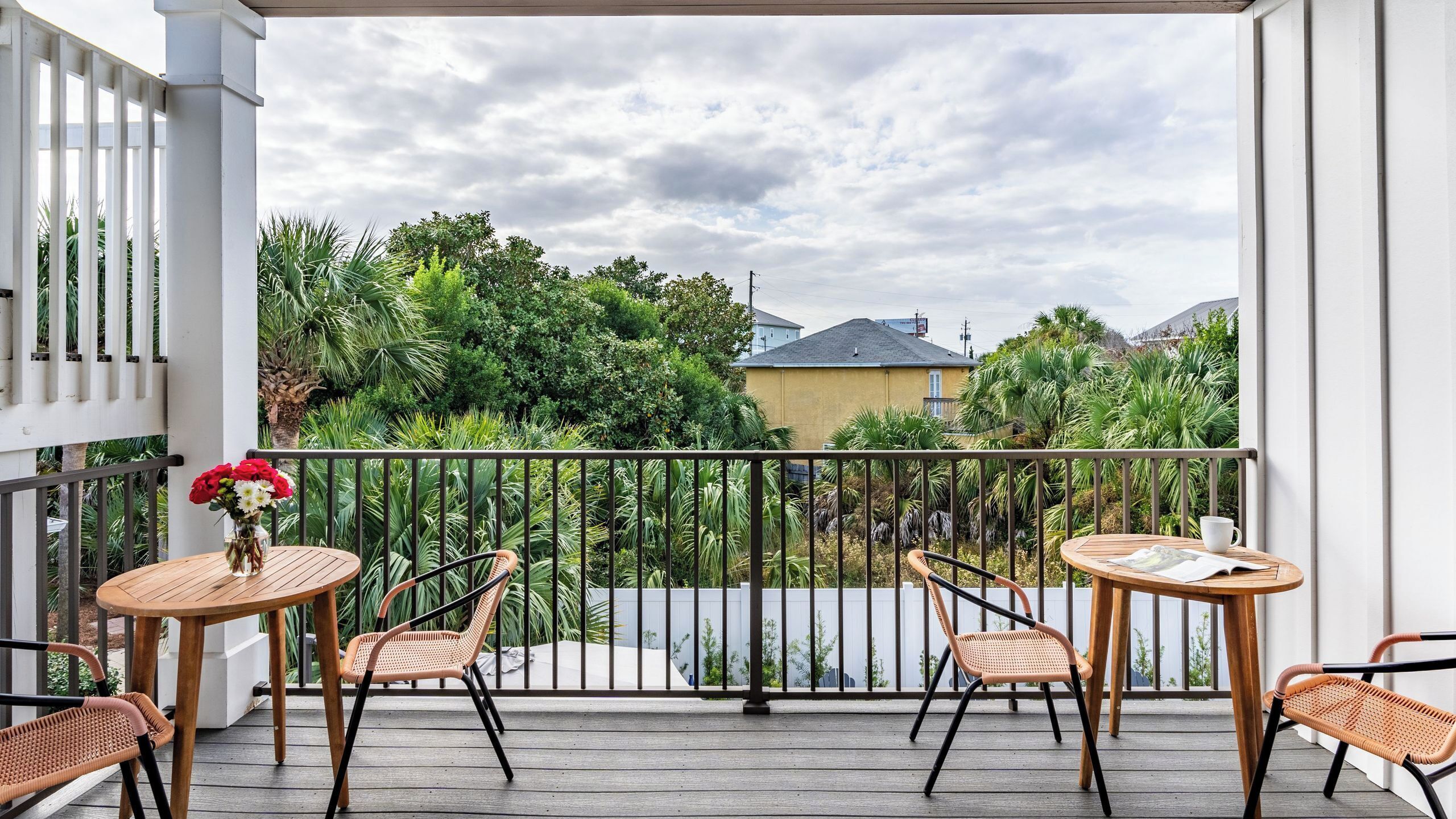 A balcony with two wooden tables and wicker chairs, a vase of flowers, and a view of greenery and houses.