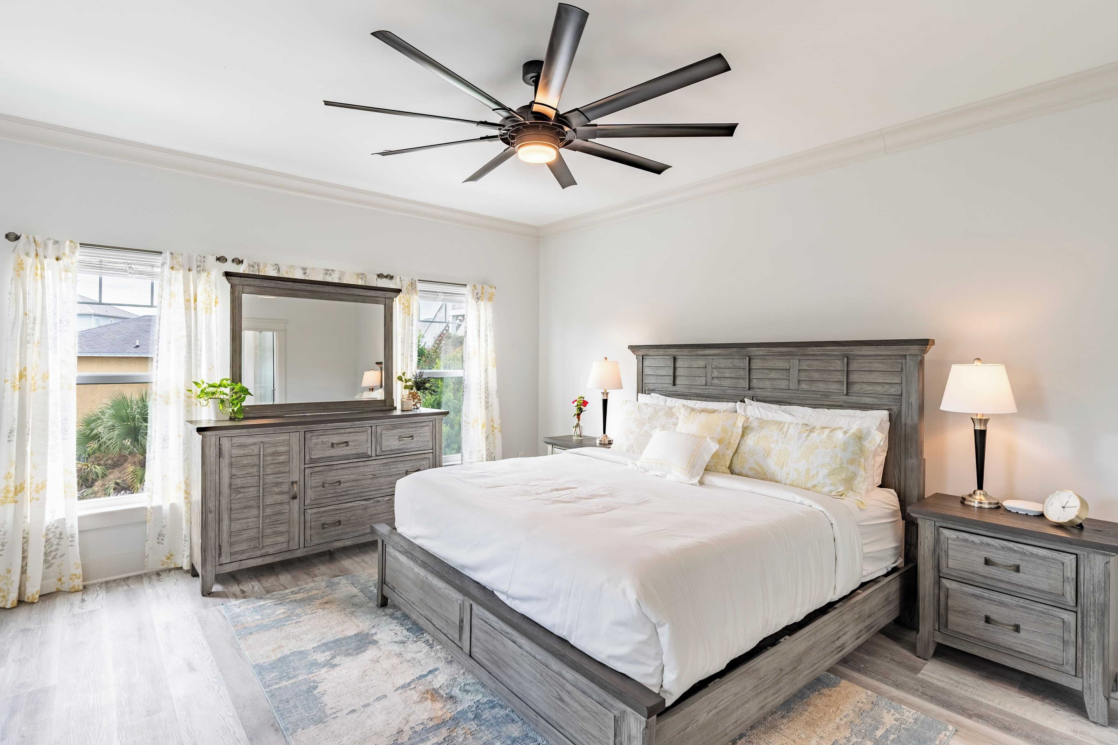 A bedroom with a large bed, a ceiling fan, a dresser, two bedside tables with lamps, and a window with curtains.