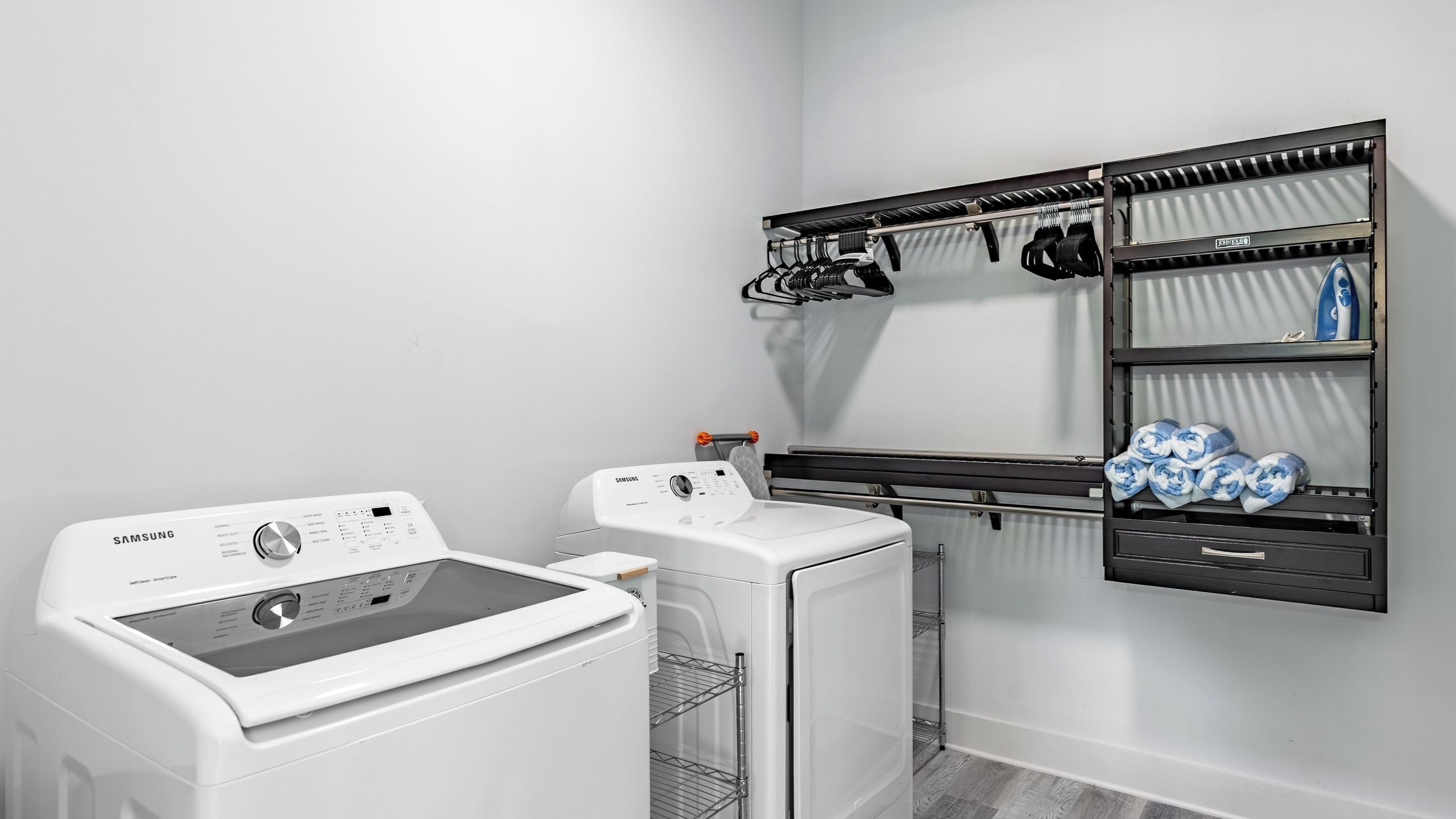 A laundry area with a Samsung washer and dryer, a metal drying rack, and a shelf with towels.