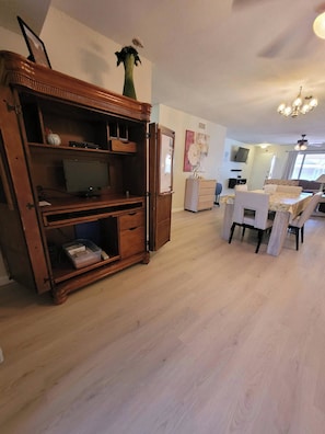 Un vaste espace de vie comprenant un meuble de rangement en bois pour les appareils multimédias, une table à manger avec des chaises, et une cuisine en arrière-plan.