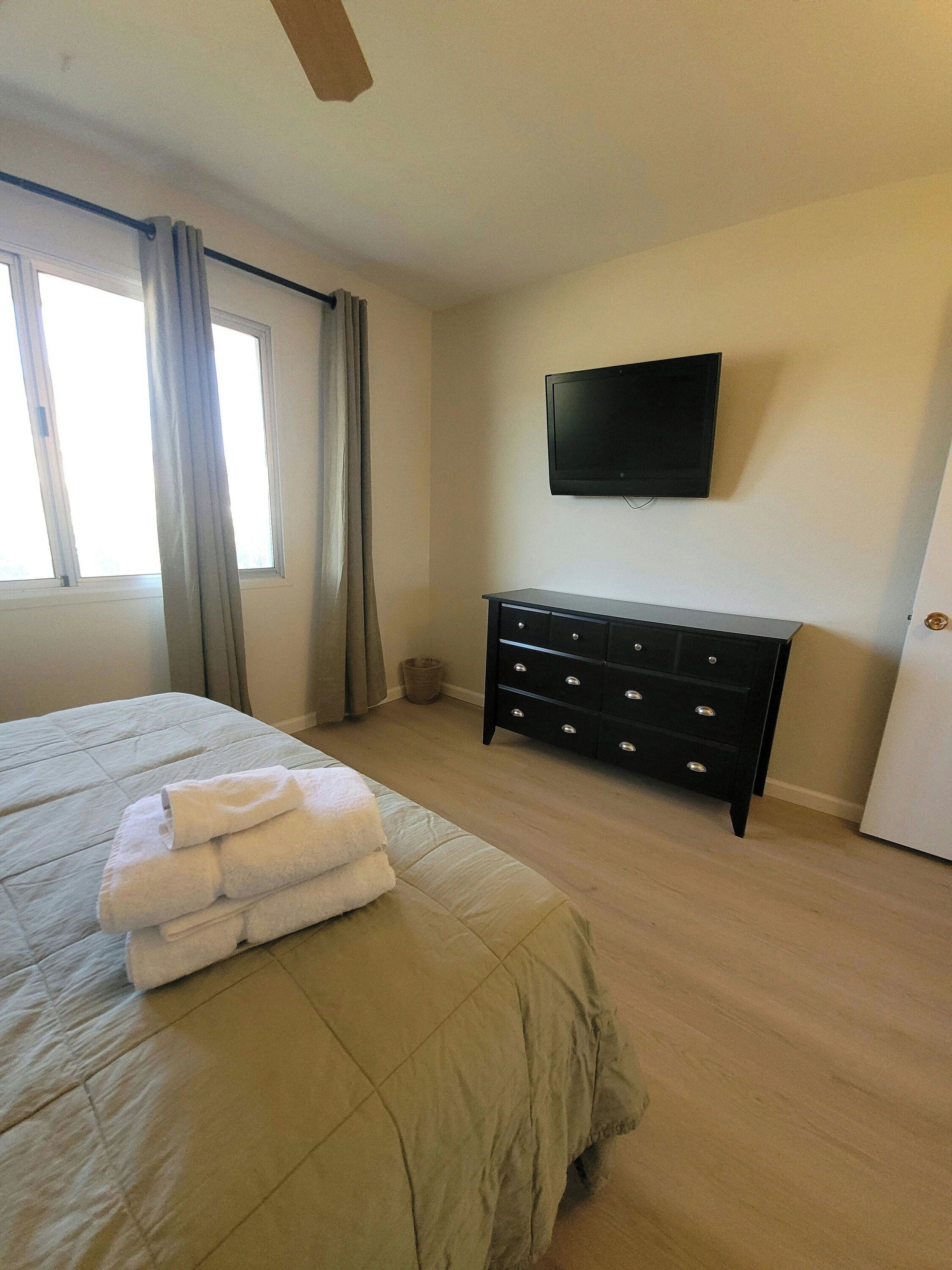 Un dormitorio con cama, cómoda, televisor y ventana con cortinas.