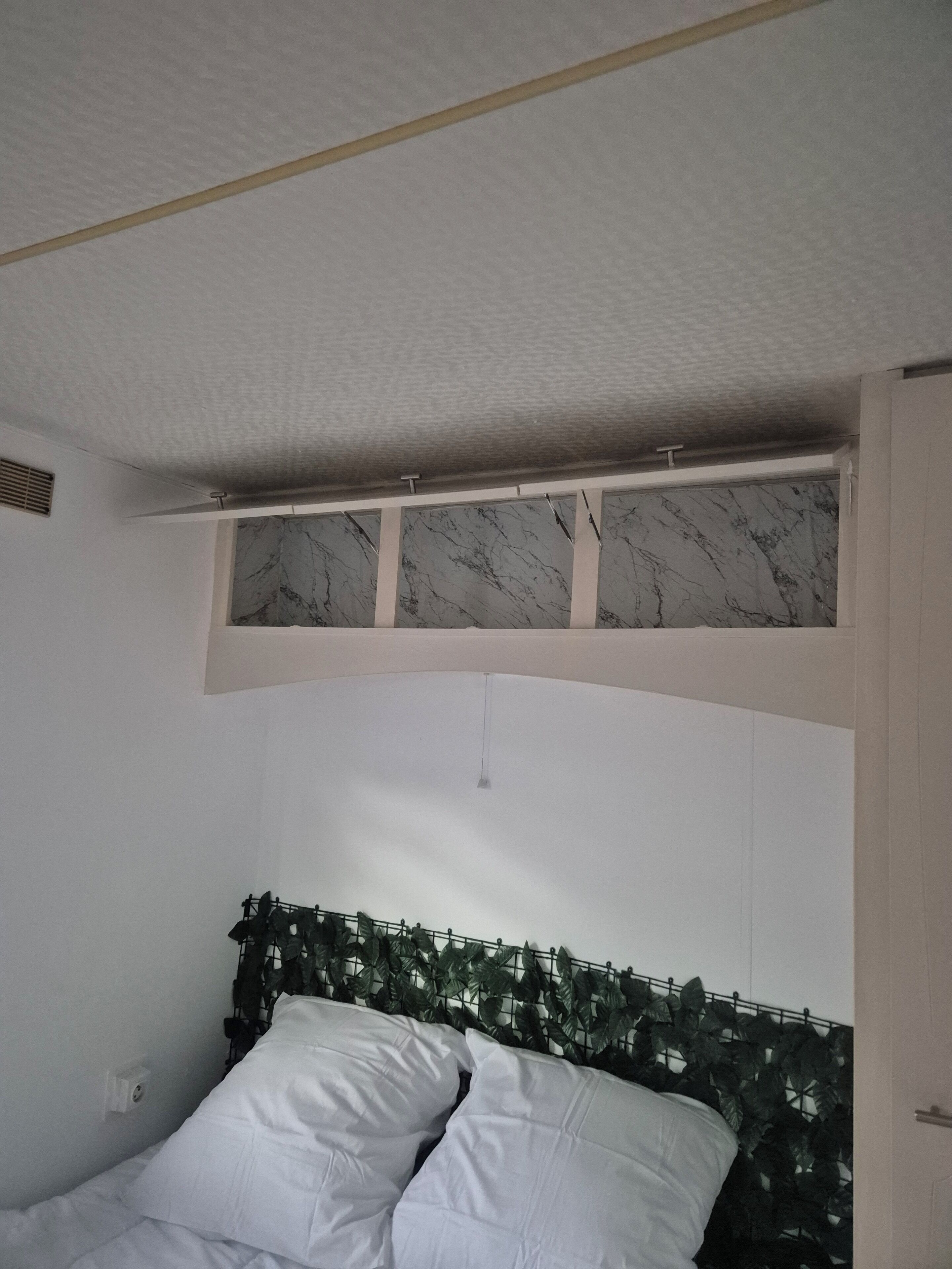 Une chambre à coucher avec un lit, des oreillers blancs, une tête de lit ornée d’un motif de feuilles, un plafond avec une bouche d’aération et un panneau mural imitant le marbre.