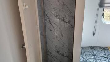 A narrow, enclosed shower area with a marble wall and a small step leading to a bed with a patterned bedspread.