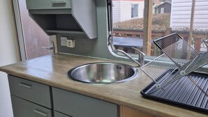 A compact kitchen with a stainless steel sink, grey cabinets, and a window overlooking a residential area. - Mobile home N°17 (Fourmies)