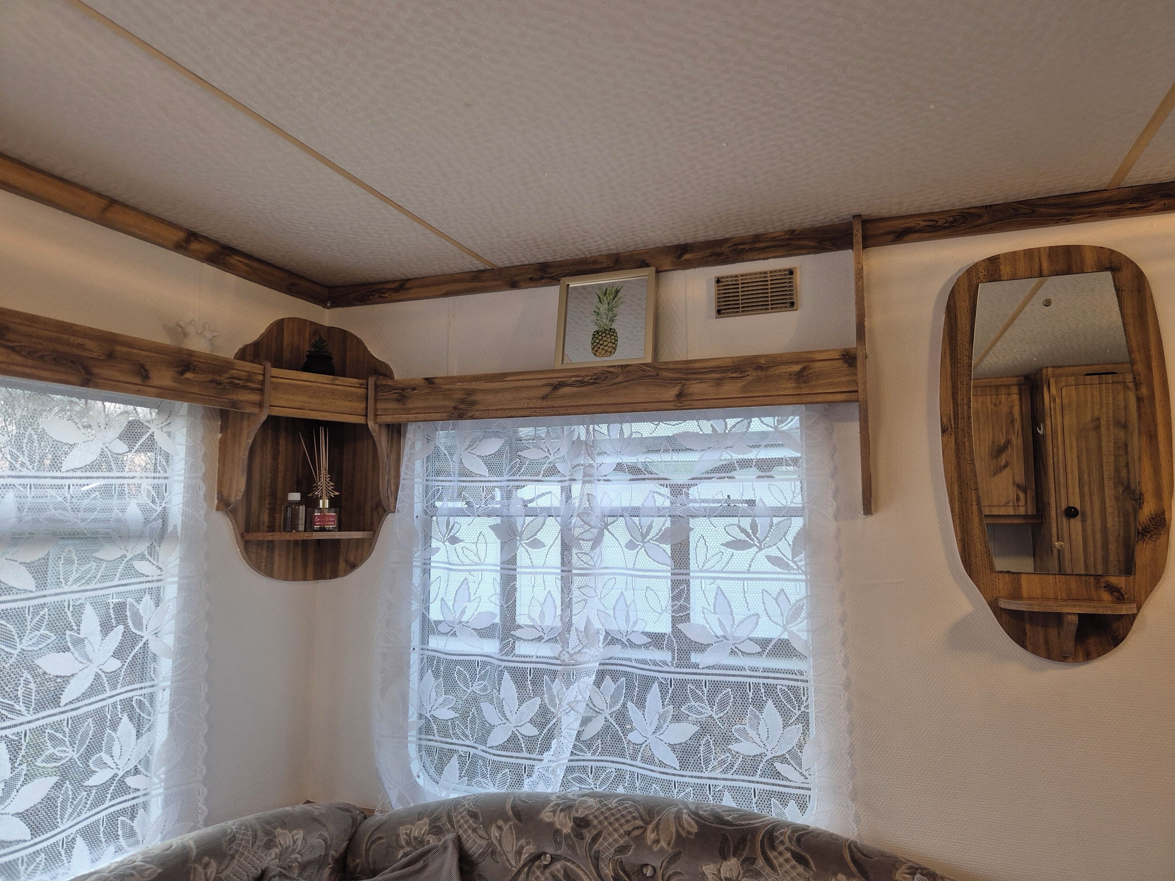 A room with a wooden ceiling, a window with lace curtains, and a mirror on the wall.