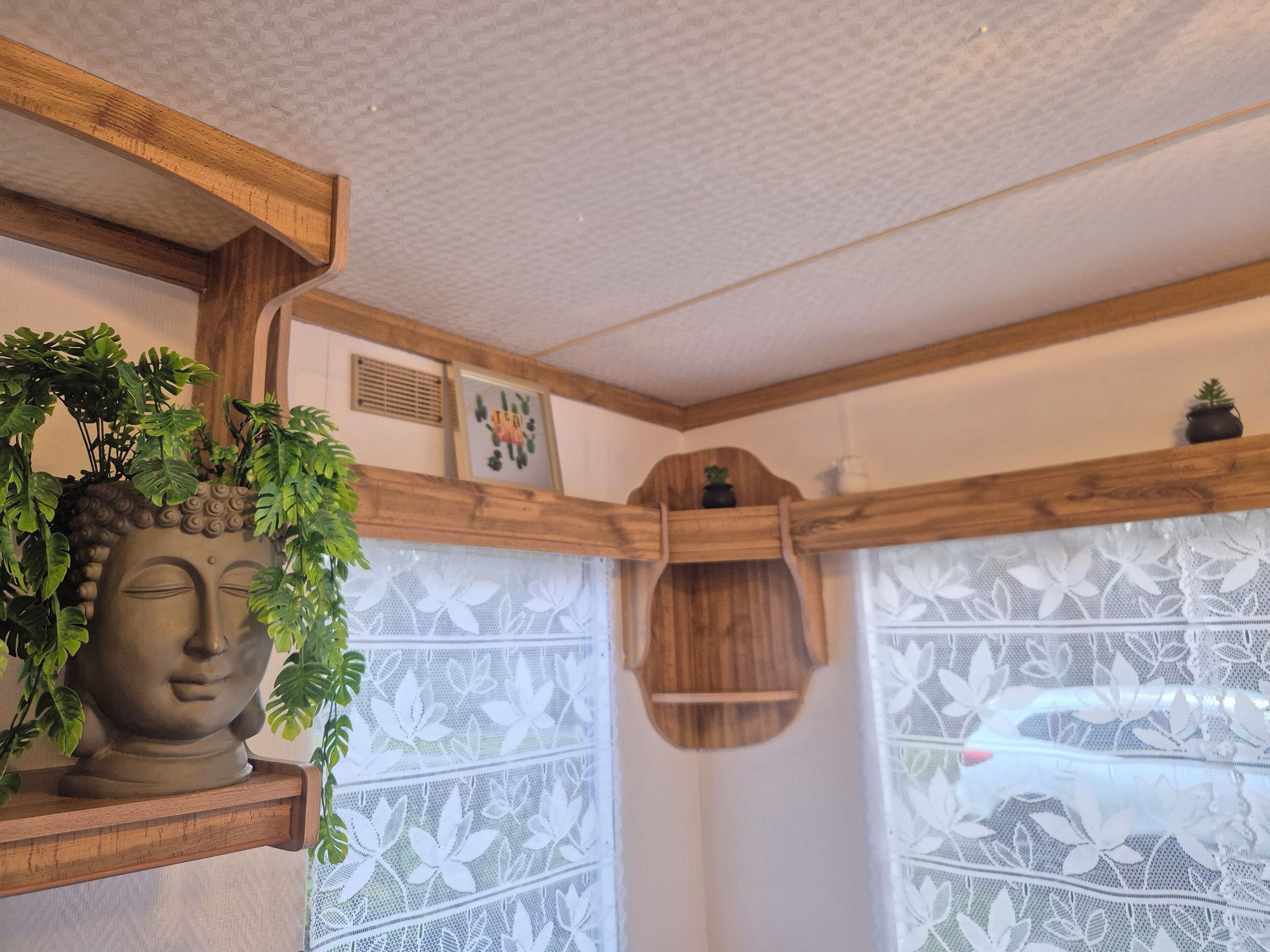 Un angolo di stanza con una mensola in legno che ospita una statua di Buddha e piante, un pannello decorativo in legno e una finestra con tende di pizzo.