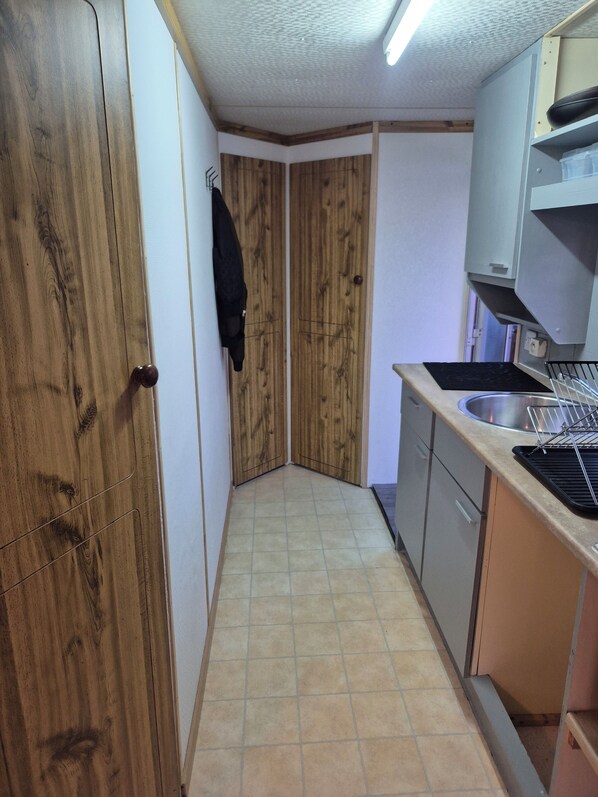 A kitchen with wooden cabinets, a sink, and a black jacket hanging on the wall. - Mobile home N°17 (Fourmies)