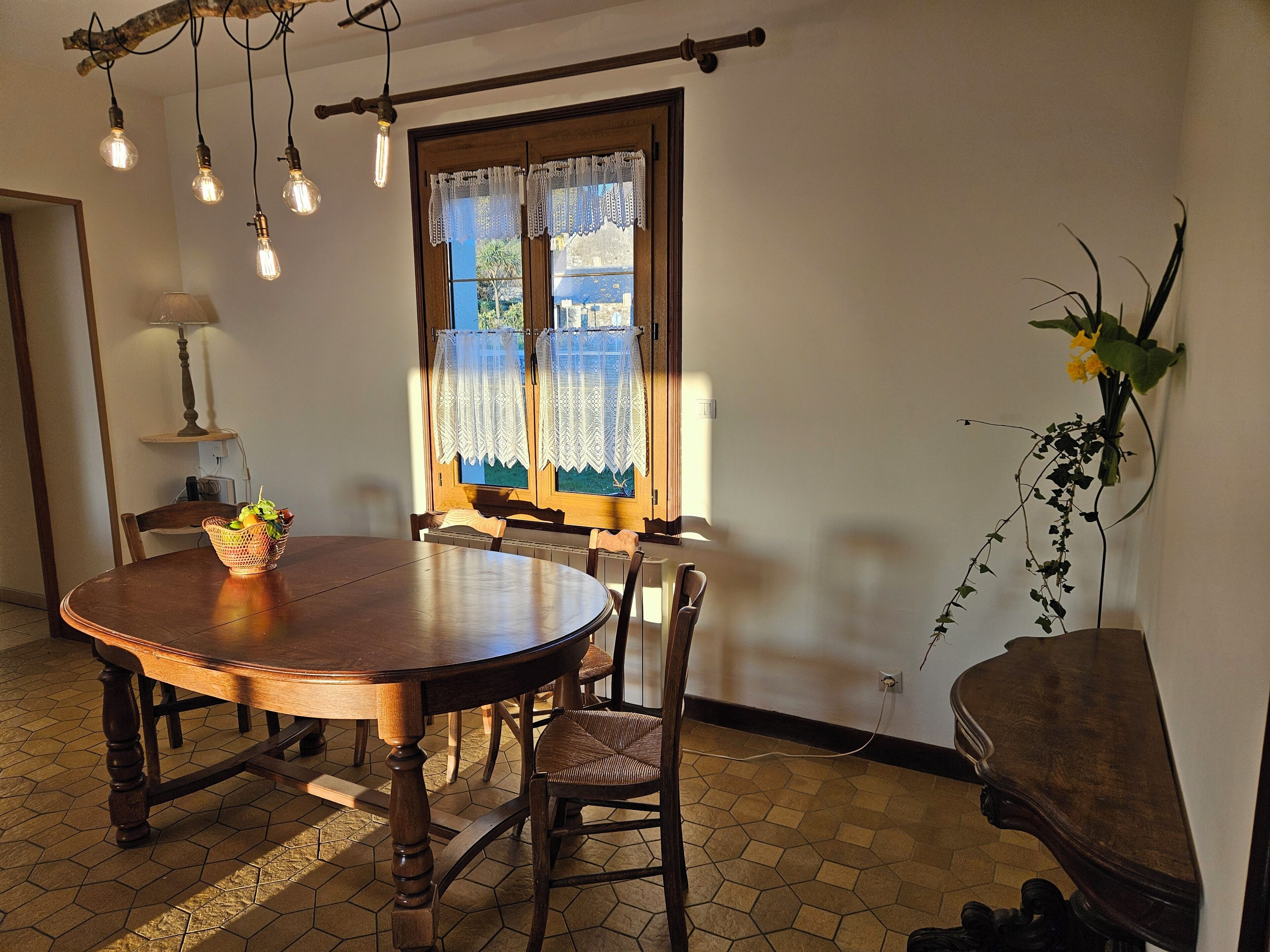 Un comedor con una mesa redonda de madera, cuatro sillas, una cesta con frutas y una ventana con cortinas de encaje.