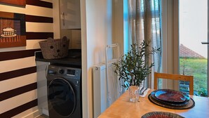 A laundry area with a washing machine, a radiator, and a table with a glass of water and plates. - Villa Noria (Kingston Bagpuize)