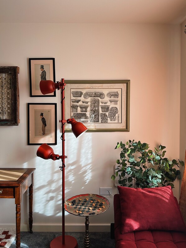 A room with a wooden floor, a red sofa, a small round table with a chessboard, a potted plant, and framed artwork on the wall.