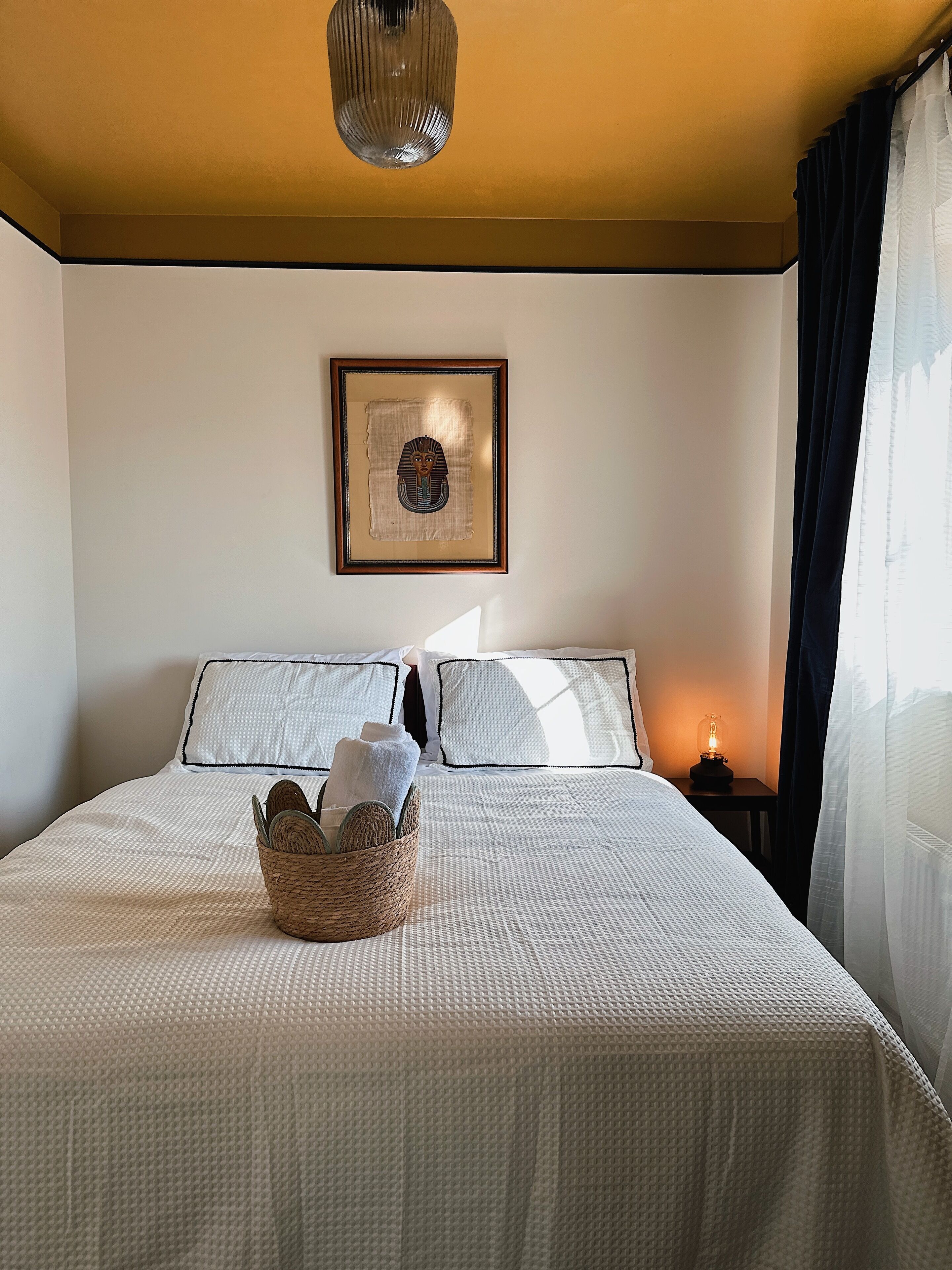 A neatly made bed with a white bedspread, a framed picture on the wall, and a small bedside table with a lamp.