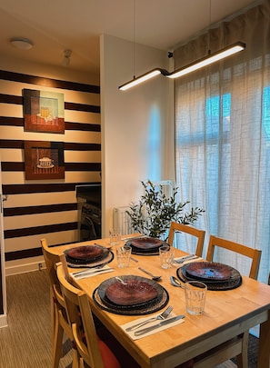 A dining area with a wooden table set for four, surrounded by chairs. There are two wall-mounted light fixtures above the table, and a potted plant on the windowsill.