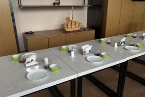 A long dining table with multiple sets of white and green plates, cups, and a ship model on a shelf above.