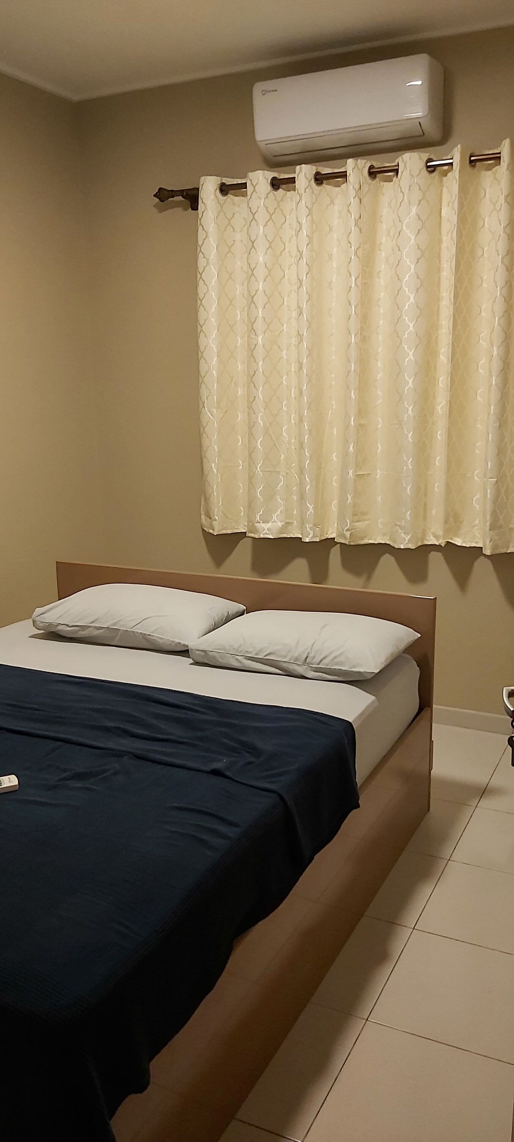 A bedroom with a bed, a window with curtains, an air conditioning unit, and a bedside table.