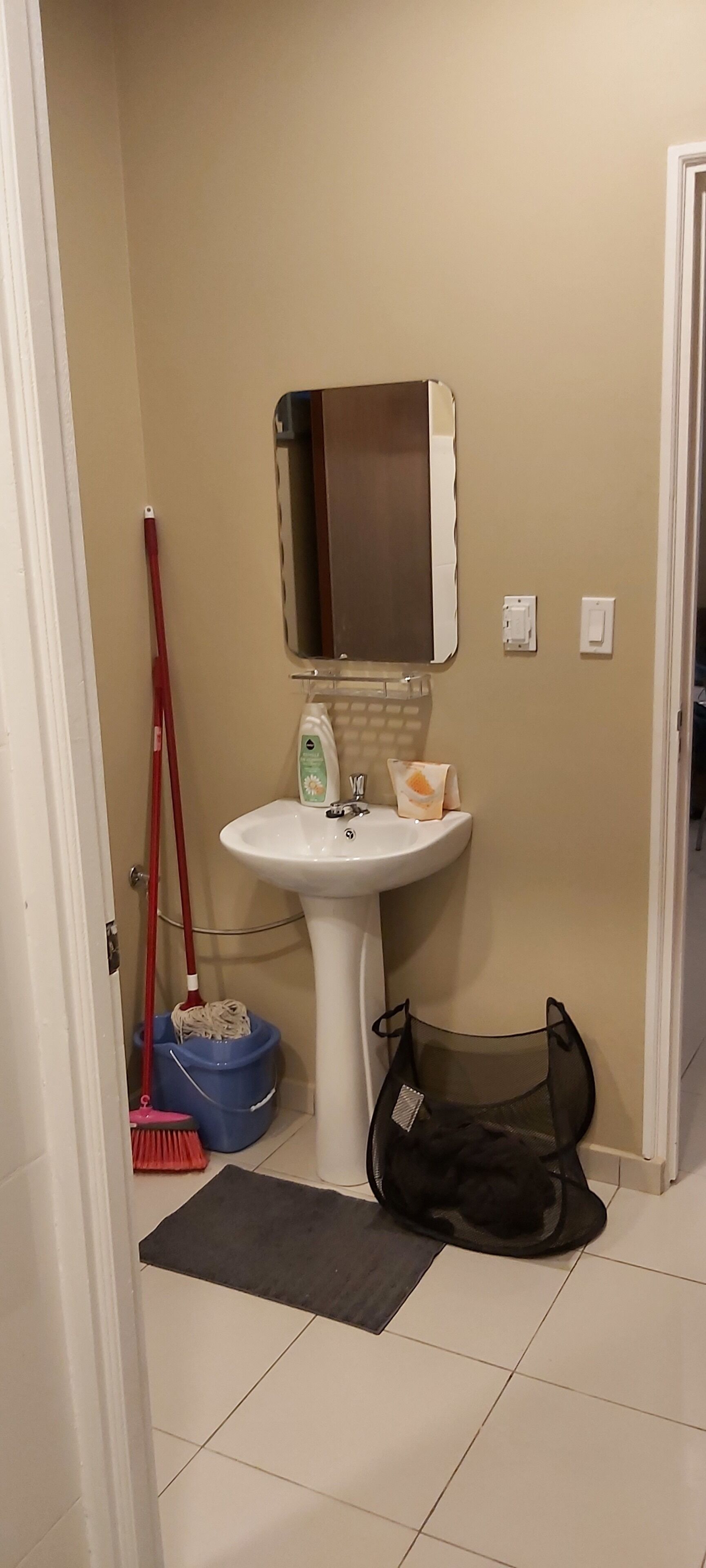 A bathroom with a white sink, a mirror, a red broom, a blue dustpan, and a black bag on the floor.