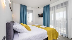 Hotellihuone, jossa on sänky, seinälle asennettu taulutelevisio ja suuret ikkunat, joissa on siniset verhot.
