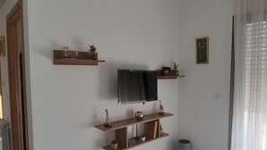Un salon doté d’un téléviseur à écran plat fixé au mur, d’étagères en bois ornées d’objets décoratifs, d’un parquet à motifs et d’une grande fenêtre agrémentée de rideaux légers.