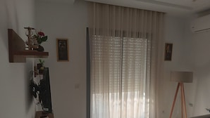 Un salon doté d’une grande fenêtre ornée de rideaux légers, d’un buffet en bois sur lequel sont posés un vase et un tableau encadré, ainsi que d’un lampadaire.