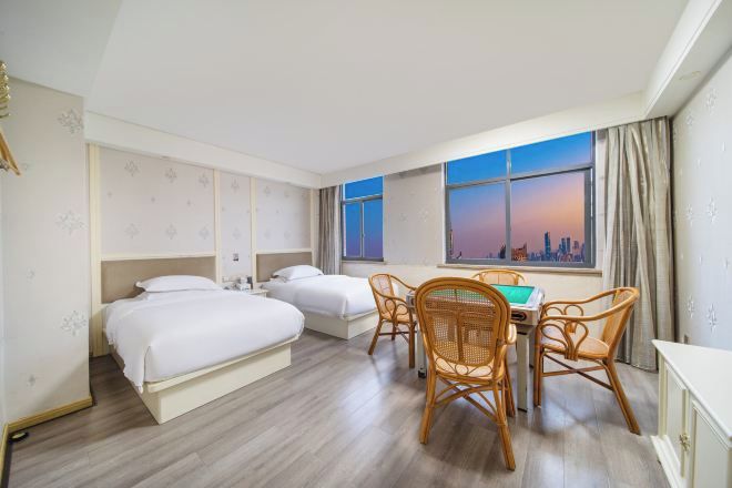 A hotel room with two beds, a dining table with chairs, and a cityscape view through the window.