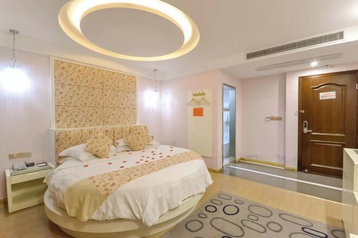 A hotel room with a round bed, a circular ceiling light, a patterned headboard, and a wooden door.
