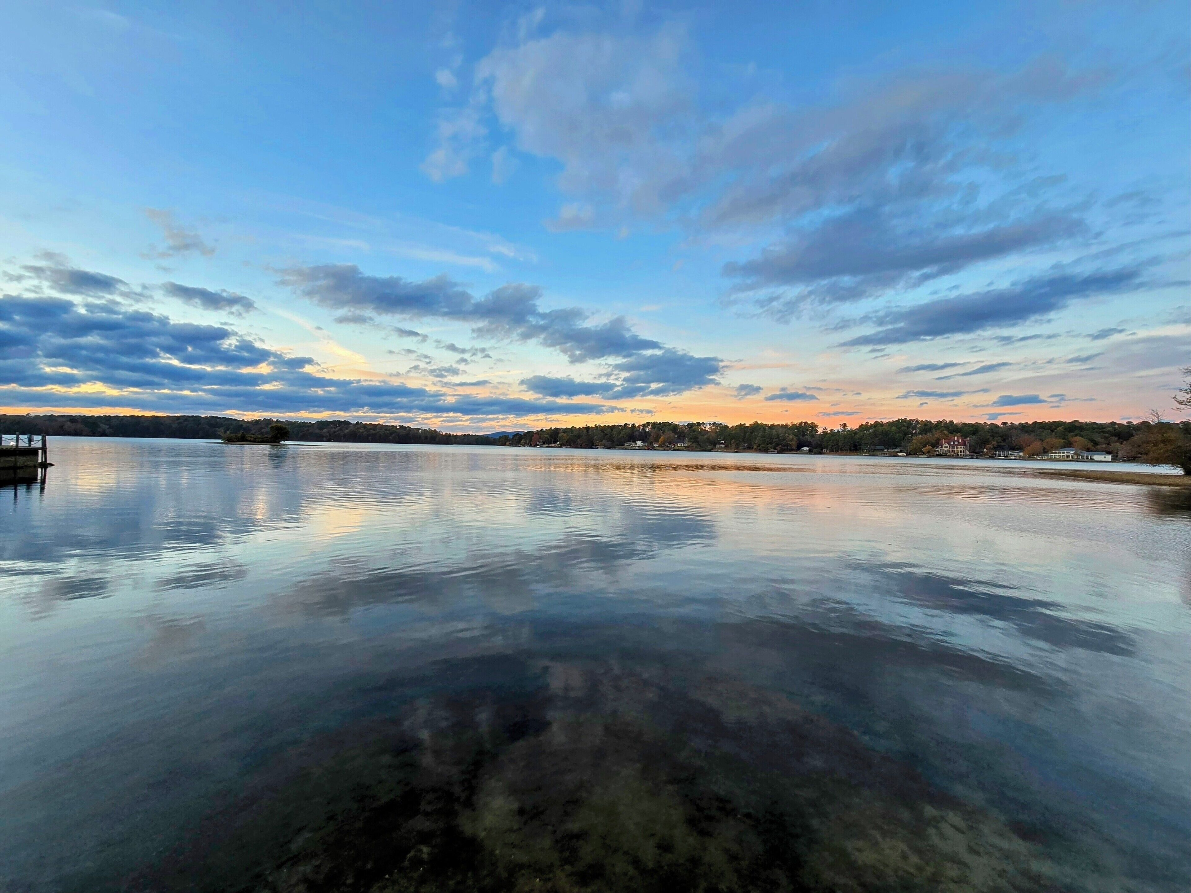 Ein ruhiger See mit einem Steg, umgeben von Bäumen und Gebäuden in der Ferne.