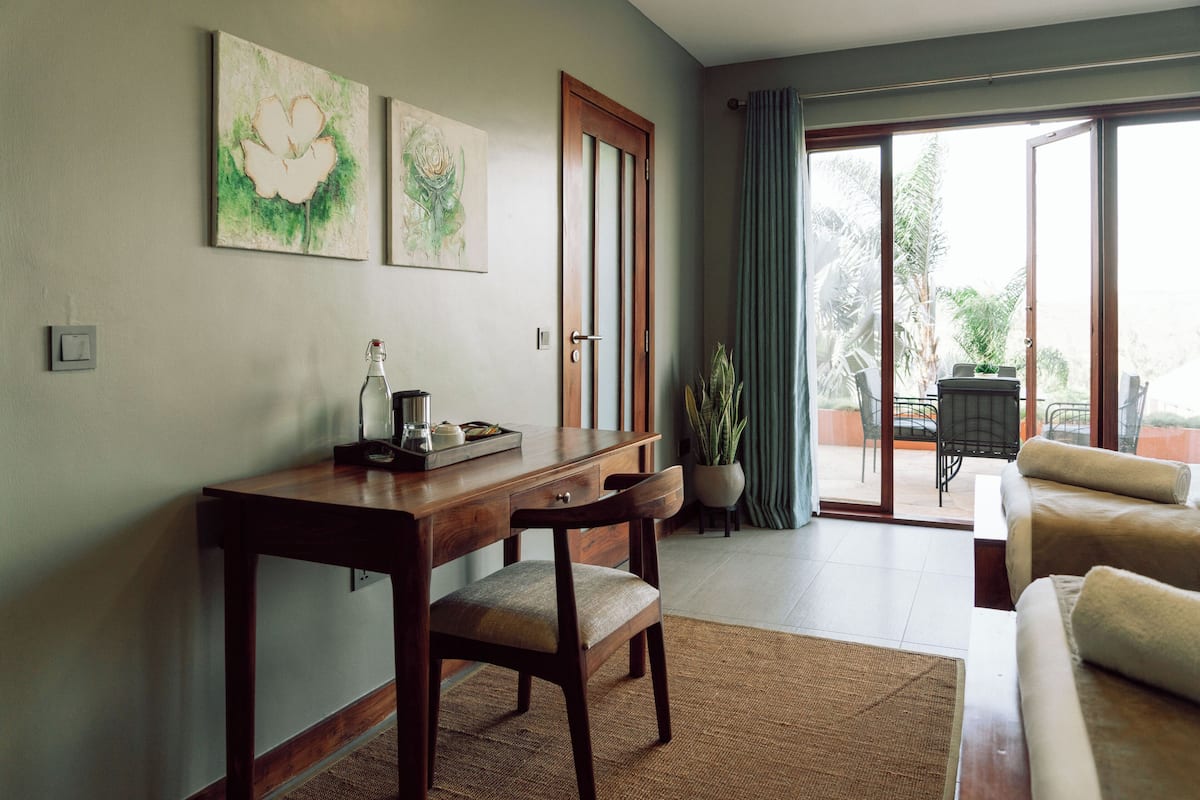A room with a wooden desk, a chair, a sofa, and a large sliding glass door leading to an outdoor area with chairs and a table.