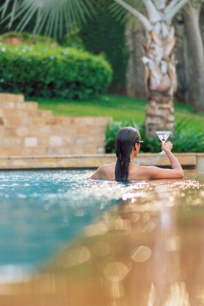 A person relaxing in a swimming pool with a drink, surrounded by greenery and stone structures. - THE SCENERY RESIDENCE (Arusha)