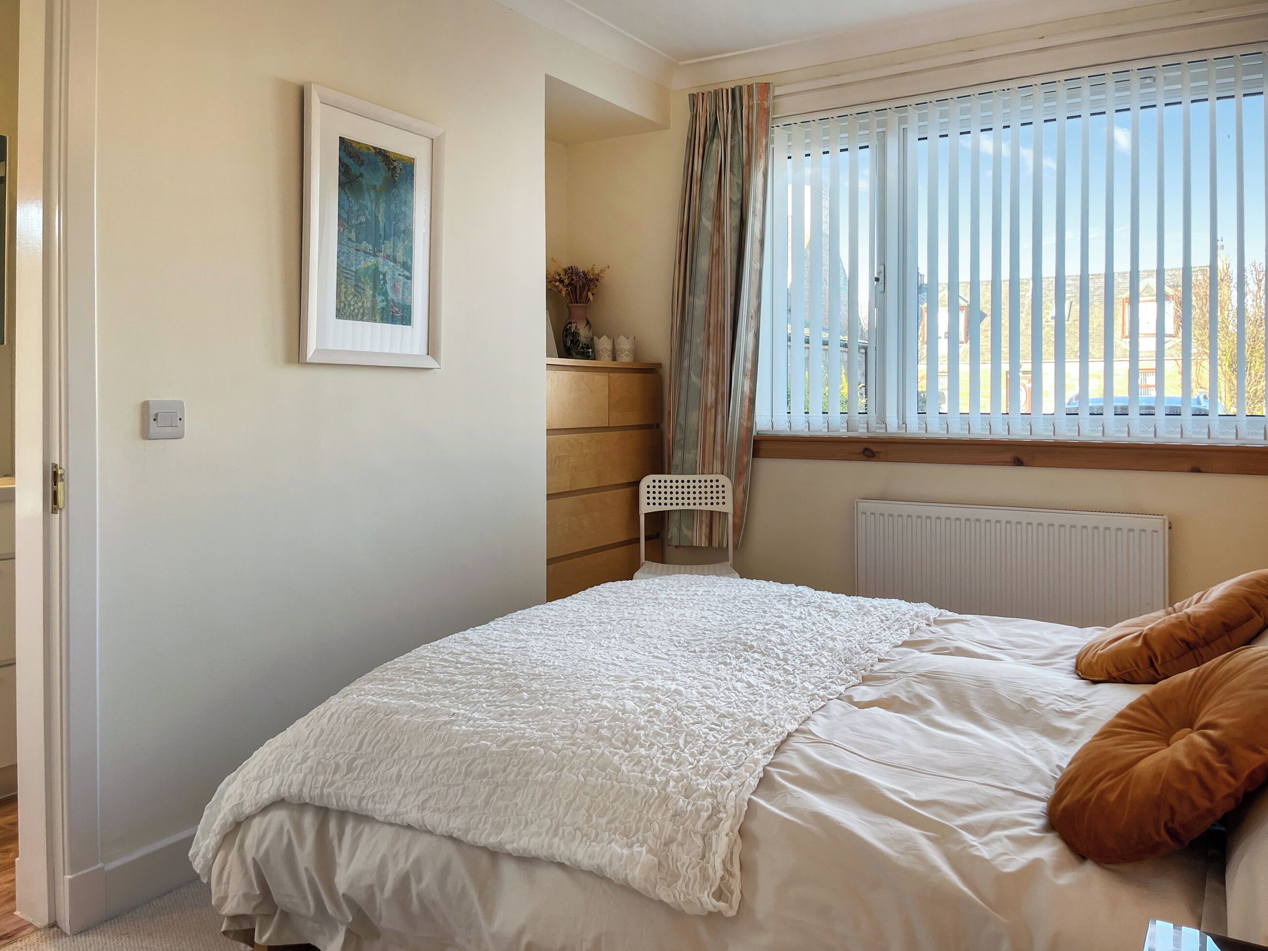Een slaapkamer met een bed, een radiator, een raam met jaloezieën, een stoel en een tafeltje met een plant.