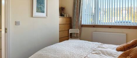 Een slaapkamer met een bed, een radiator, een raam met jaloezieën, een stoel en een tafeltje met een plant.