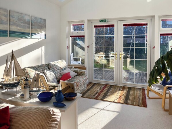 A sunlit living room with a patterned sofa, a small sailboat model, and a view through glass doors to an outdoor deck.