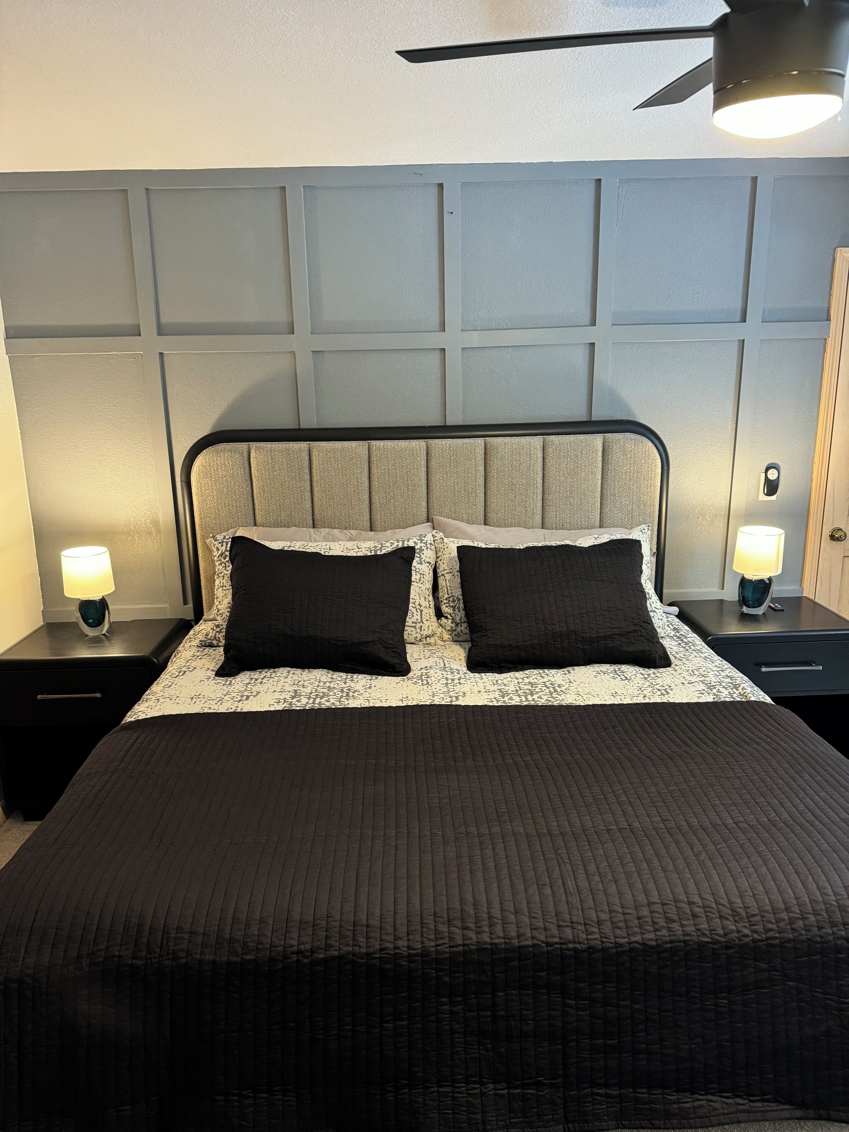 Una cama bien hecha con colcha oscura, dos almohadas negras y un cabecero con diseño de panel texturizado. Hay dos mesitas de noche con lámparas y un ventilador de techo sobre la cama.