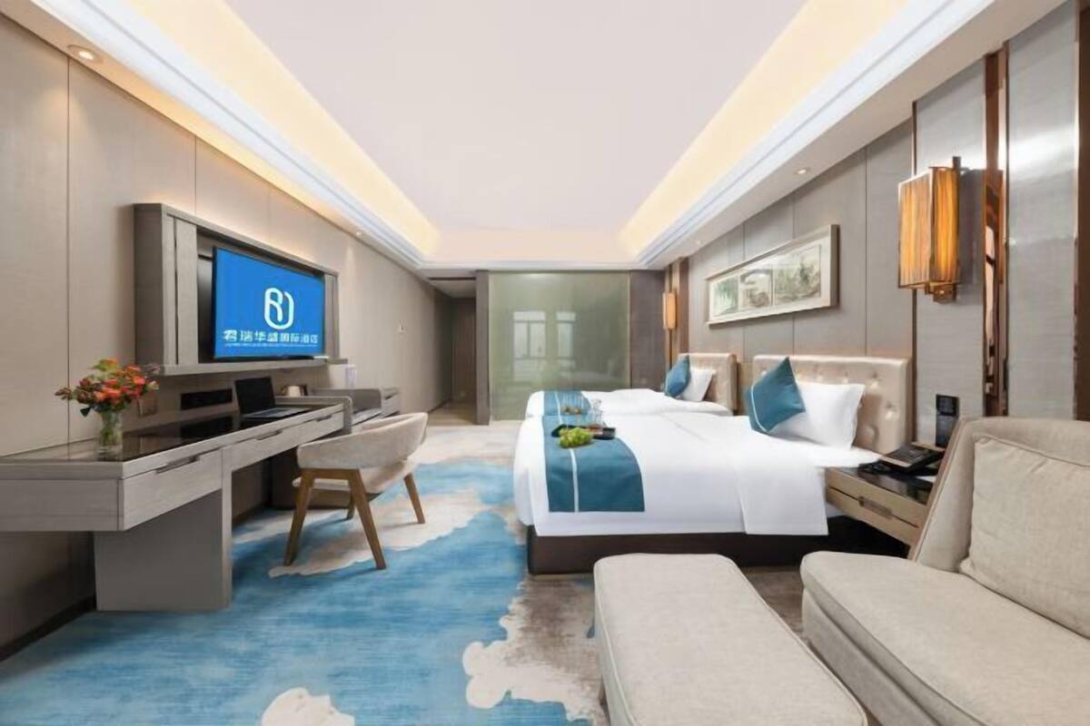 A modern hotel room with a bed, a desk, a chair, a sofa, a TV, and a wall-mounted lamp.