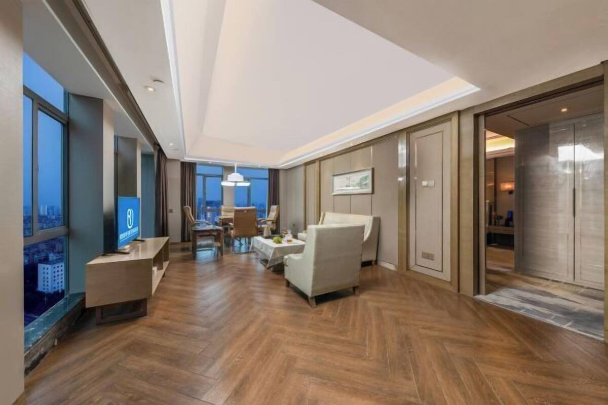 A spacious hotel lobby with a wooden floor, a seating area, and a large window offering a city view.
