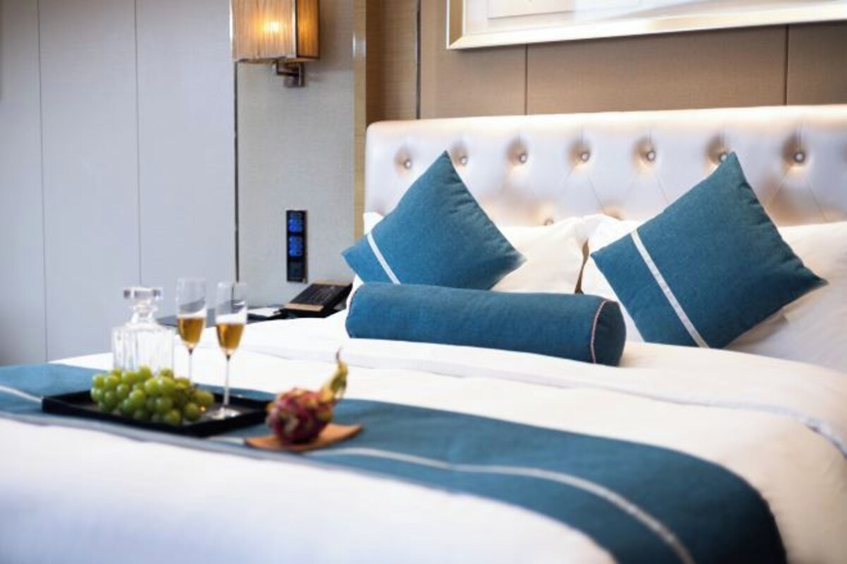Een netjes opgemaakt bed met blauwe kussens, een dienblad met druiven en glazen wijn, en een nachtkastje met een lamp en telefoon.