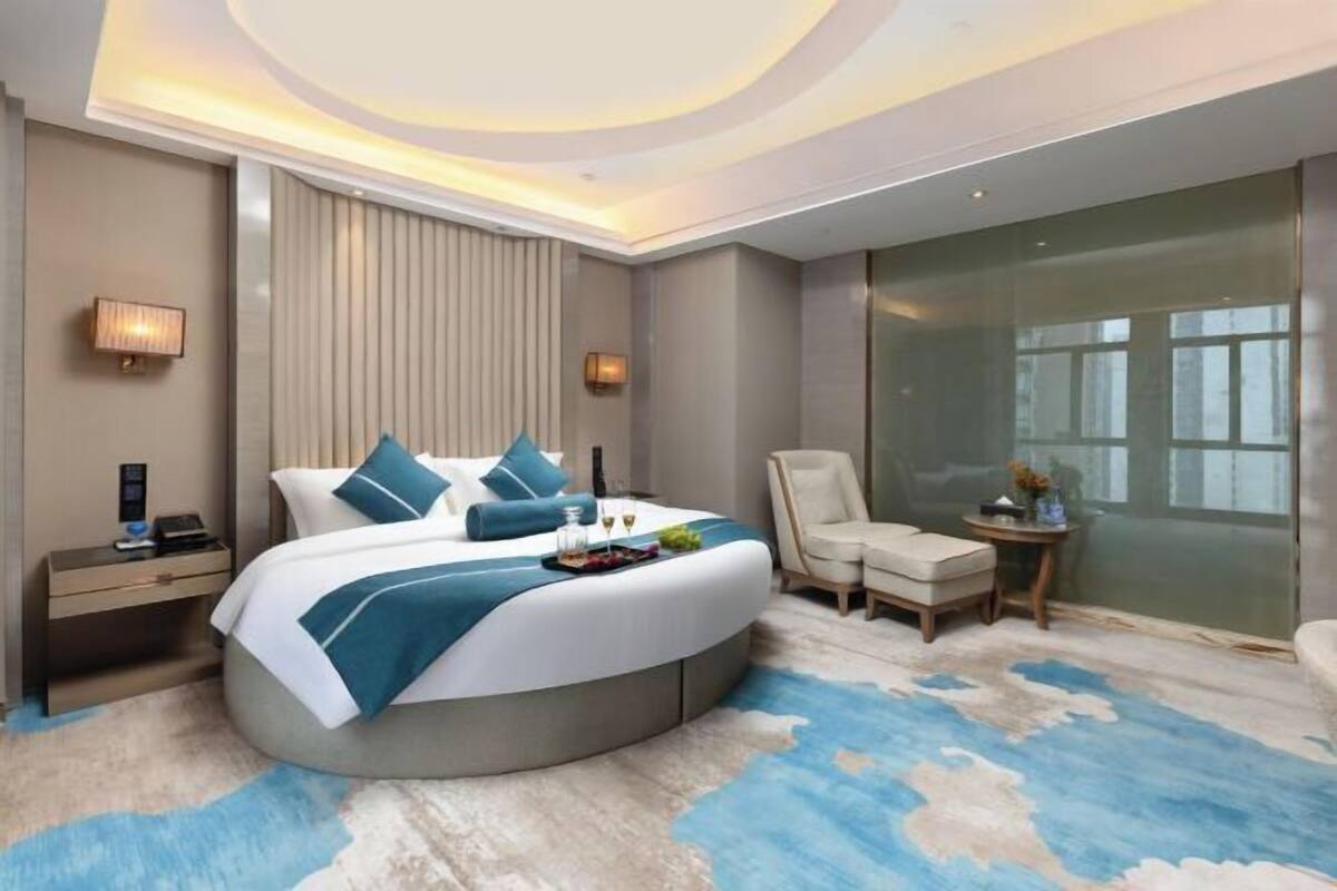 A modern hotel room with a curved bed, a seating area, a glass partition, and a large window.
