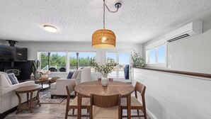 A spacious living area with a round wooden table and chairs, a fireplace, and large windows offering a view of greenery. - Sunset Shores - Ocean Front Family Retreat HOT TUB  (Ocean Park)