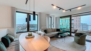 Un salon moderne avec une table à manger en bois, un canapé et un fauteuil. De grandes fenêtres offrent une vue sur la ville.