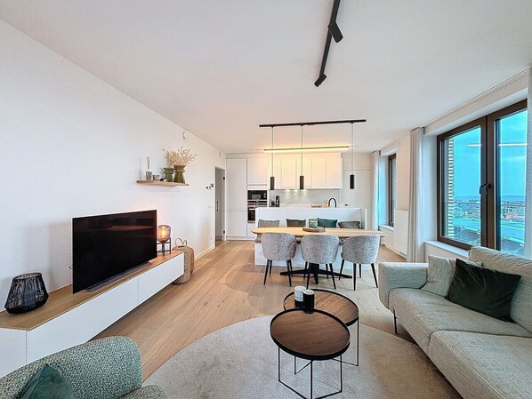 A modern living room with a flat-screen TV, a dining area with a table and chairs, and a large window with a city view.
