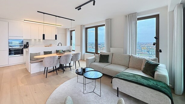 Un appartement moderne comprenant une cuisine, un coin repas et un salon. Il est doté de grandes fenêtres, d’un canapé et d’une table basse.