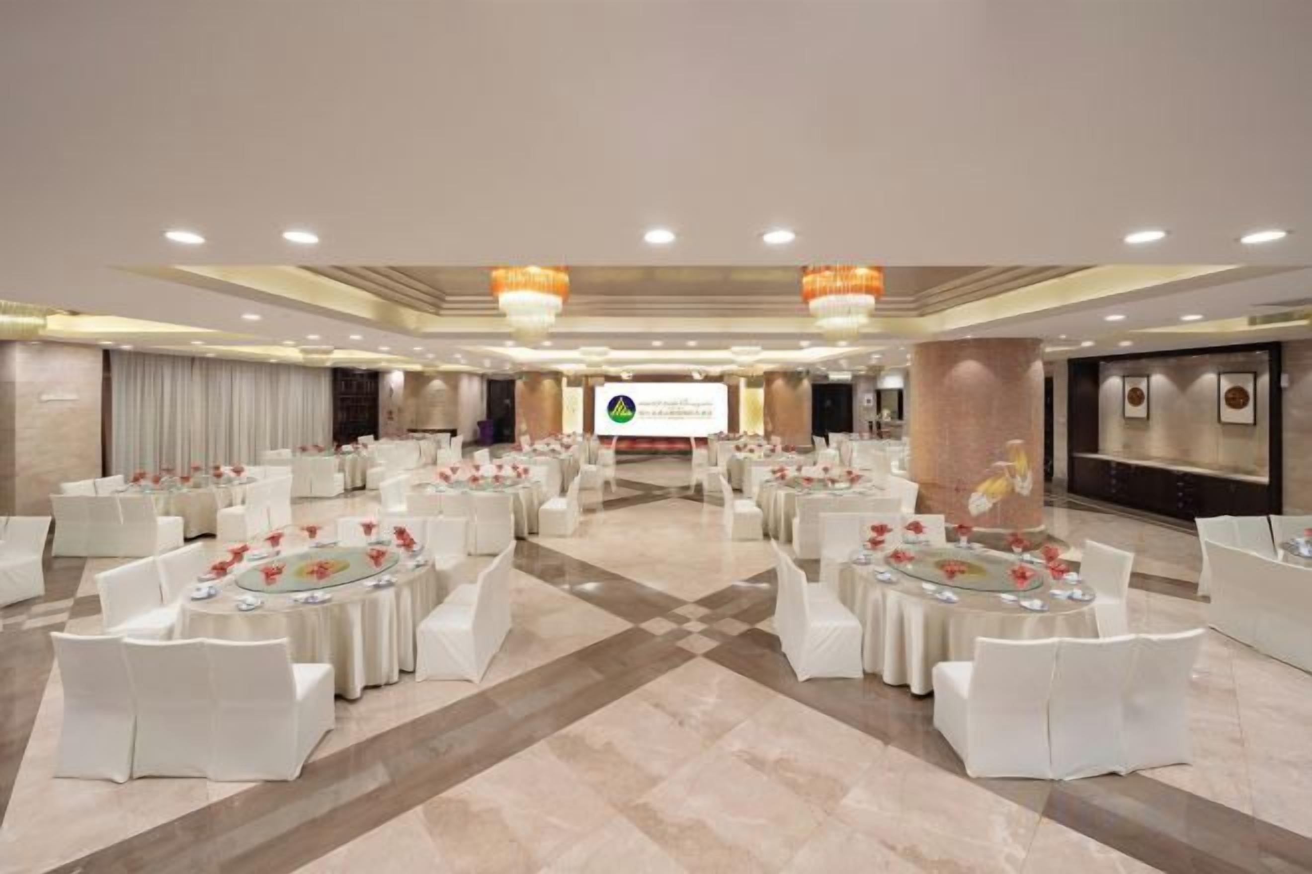 Een ruime congreszaal met ronde tafels en witte stoelen, een podium met een presentatiescherm, en een modern interieur.