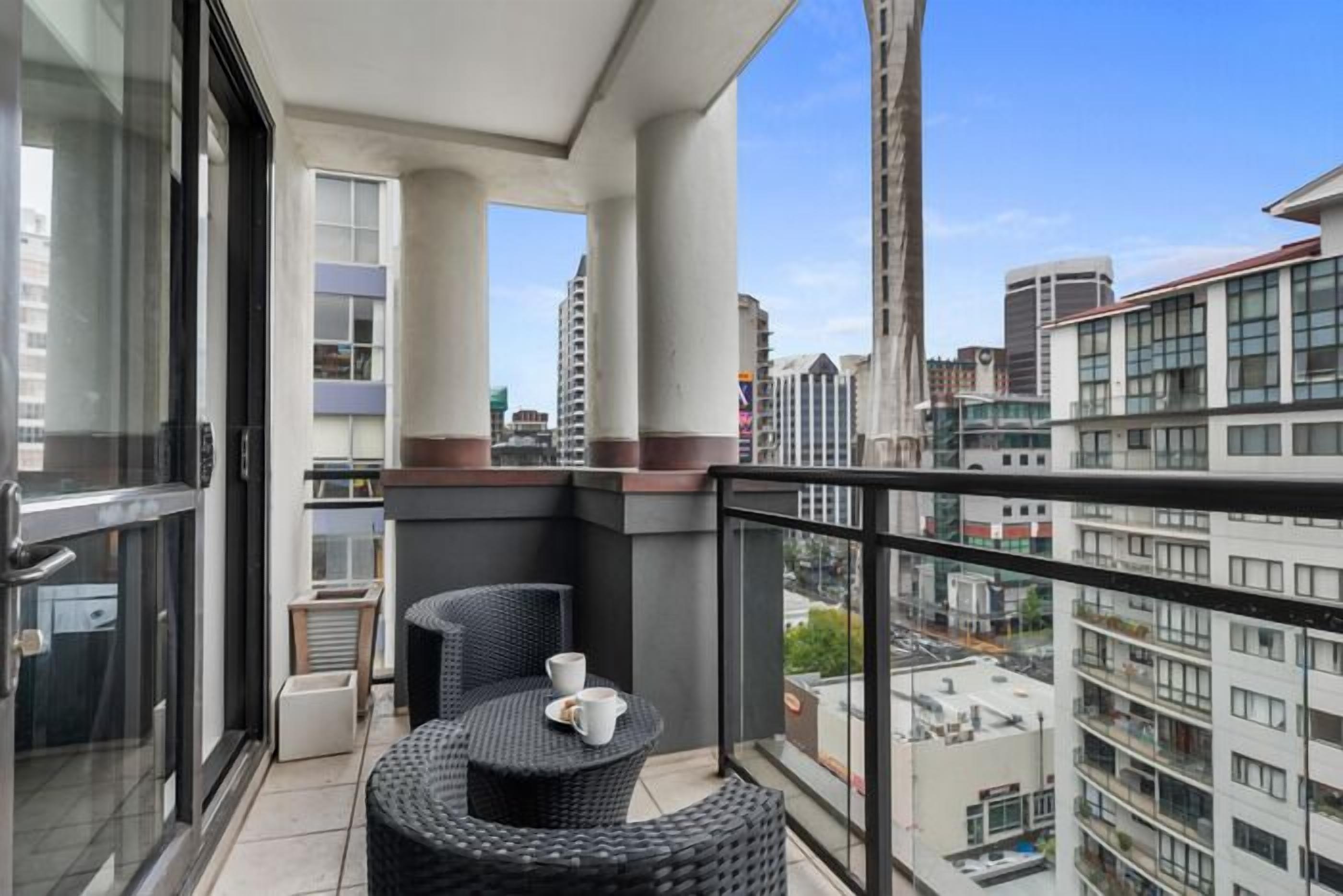 Ein Balkon mit einem Korbstuhl-Set, einem Blumentopf und Blick auf umliegende Gebäude.