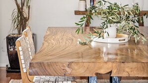 Une table à manger au plateau en bois, entourée de chaises dont les dossiers sont en osier. Un pot de fleurs avec des feuilles vertes est posé sur la table.
