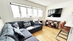 Een moderne woonkamer met een hoekbank, een houten salontafel, een aan de muur bevestigde televisie en een eethoek met een stoel en tafel.