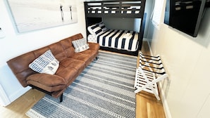 Een kleine kamer met een bruine bank, een stapelbed met gestreepte lakens, een kamerkleed met een patroon en een televisie aan de muur.